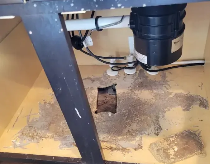 A swollen sink cabinet floor with an access hole cut beneath the plumbing