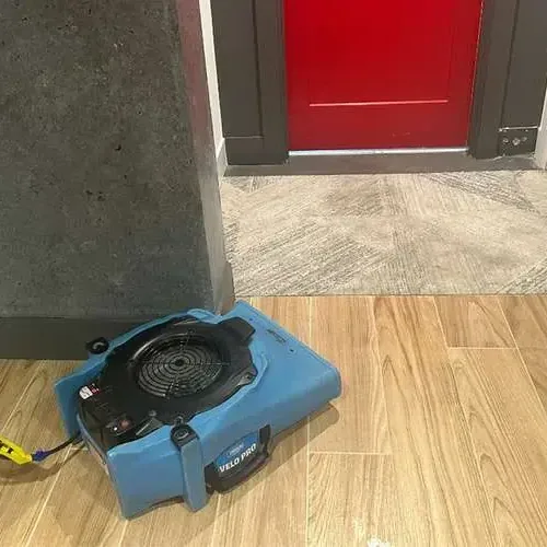 An air mover drying a wood-look tile floor in a room beside an open doorway
